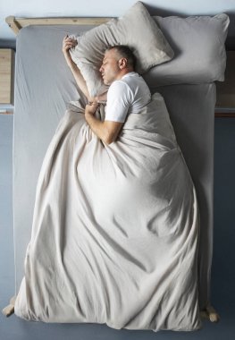 Orta yaşlı bir adamın huzur içinde uyuduğunu, yastığı kucakladığını, battaniyeyi tuttuğunu, yatakta yattığını, evde şekerleme yaptığını.