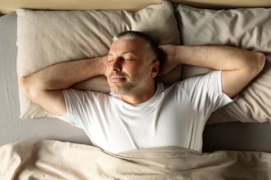 Huzurlu orta yaşlı bir adamın yatakta gözleri kapalı, el ele tutuşmuş, battaniyeyle örtülü olması. Sağlıklı dinlenme kavramı