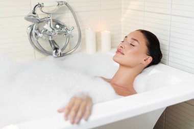 Rahatlamış genç Avrupalı bayan köpükle banyo yapıyor, banyoda küvette uzanıyor, evde vücut bakımının keyfini çıkarıyor.