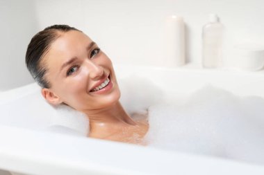 Rahatlama ve banyo konsepti. Mutlu Avrupalı kadın portresi köpüklü banyo yapıyor, kameraya gülümsüyor, banyo küvetinde suda dinleniyor.