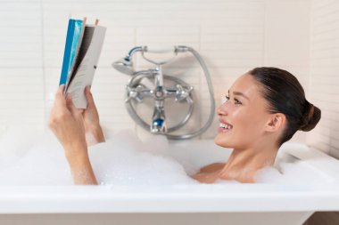 Mutlu genç kadın köpüklü banyoda uzanıyor ve dergi okuyor evdeki küvette rahatlatıcı atmosferde, yan bakışta.