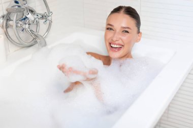 Mutlu kadın banyo yapıyor ve eğleniyor, köpük üflüyor ve gülüyor, küvette sabun köpüğü dolu bir küvette oturuyor,
