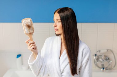 Saç dökülme problemi. Üzgün kadın saçlarıyla dolu tarağa bakıyor, alopecia ya da vitamin eksikliği çekiyor, banyonun içinde duruyor.