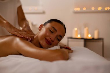 Rahatlamış ve tatmin olmuş genç bir bayan sırt masajı yaptırıyor. Masör omuzlarına masaj yaparken gözleri kapalı yatıyor. Hoş bir atmosfer ve mumlar.