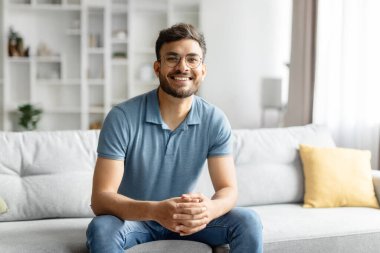 Yakışıklı Hintli bir adamın portresi evde tek başına dinleniyor, rahat bir oturma odasında kanepede oturuyor ve kameraya gülümsüyor, neşeli Hintli adam günlük kıyafetli, boş alan.