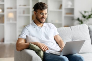 Pozitif Hintli adam ev içi koltukta dizüstü bilgisayar kullanarak çalışıyor, adam internette sörf yapıyor, modern teknolojiler kullanıyor, serbest alan kullanıyor.