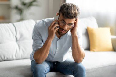 Kafası karışmış Hintli adam cep telefonuyla konuşuyor, kafaya dokunuyor, evde rahatsız edici cep telefonu konuşmaları yapıyor, oturma odasındaki koltukta oturuyor.