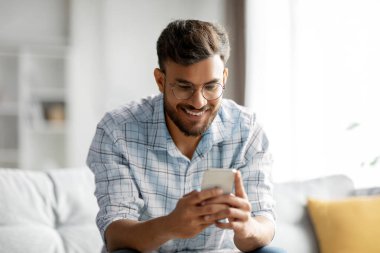 Genç gülümseyen Hintli adam cep telefonu tutuyor, SMS yazıyor ya da sosyal medyada kayıyor, açık oturma odasındaki koltukta oturuyor, boş yer var.