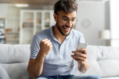 Evet, harika haberlerim var. Aşırı neşeli Hintli adam akıllı telefon tutuyor ve sıkılmış yumruğunu sallıyor, evdeki rahat koltukta oturuyor, boş yer var.