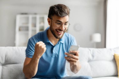 Evet, harika haberlerim var. Aşırı neşeli Hintli adam akıllı telefon tutuyor ve sıkılmış yumruğunu sallıyor, evdeki rahat koltukta oturuyor, boş yer var.