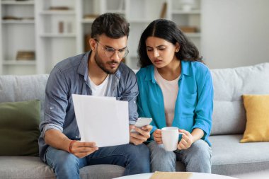 Evli Hintli çift gazete okuyor, harcamalarını hesaplıyor, birlikte bütçe planlıyorlar, cep telefonu ve muhasebe vergileri kullanıyorlar, evdeki kanepede oturuyorlar.
