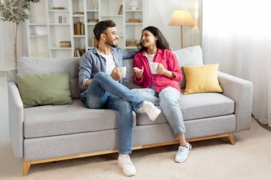Mutlu Hintli çift rahatlıyor ve kahve içiyor, evdeki kanepede oturuyor, modern oturma odasında sıcak içeceklerle dinlenirken sohbet ediyorlar,