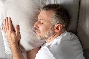 Orta yaşlı Avrupalı bir adam kulağında turuncu kulak tıkacı olan bir yatakta uyursa tüm gece boyunca huzur ve sessizlik verir. Yatak odasındaki yastıktaki adam yüksek kalite fotoğraf.