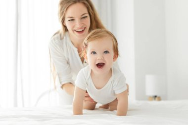 Küçük kızıyla evde yatakta oynarken gülümseyen mutlu Avrupalı bir anne, kameraya sürünen bir çocuk. Aile zamanı ve çocuk bakımı kavramı