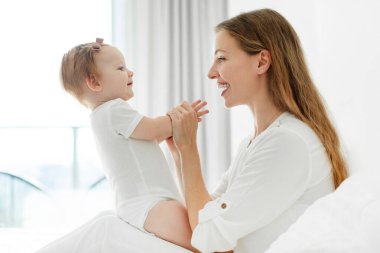 Sevgi dolu genç bir anne yatak odasında kızıyla oynuyor, annesi çocuğuna bakıyor ve gülümsüyor.