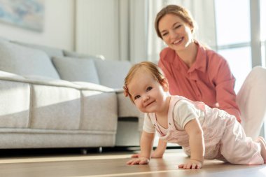 Genç Avrupalı bir kadın, güzel kızının evde dört ayak üstünde sürünmesini izliyor ve gülümsüyor, boşluğu kopyalıyor.