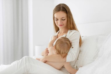 Güzel anne bebeğini emziriyor, yatakta oturuyor ve küçük kızına sarılıyor, çocukları sağlıklı bir şekilde emziriyor.