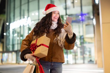 Hediye kutusu ve alışveriş torbalarıyla mutlu genç bir kadın akıllı telefon ekranına bakıyor, açık havada alışveriş merkezinin önünde duruyor.