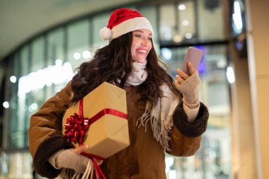 Noel Baba şapkalı heyecanlı bir kadın alışveriş merkezinin yakınında yürüyor. Hediye kutusunu taşırken ve cep telefonu kullanırken daha fazla Noel hediyesi arıyor.