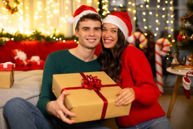 Romantik Avrupalı eşler hediye paketini tutuyor ve kameraya gülümsüyor, Noel tatillerini evde kutluyorlar.