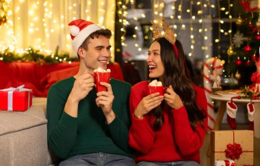 Mutlu Avrupalı çift ellerinde kakaoyla kahve bardakları tutuyorlar ve evde Noel arifesinin tadını çıkarıyorlar, birbirlerine bakıp gülümsüyorlar.