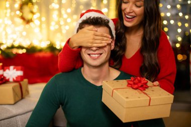 Mutlu Avrupalı bayan kocaya gözlerini kapar ve dekore edilmiş oturma odasının içinde parıldayan çelenklerle birlikte yeni yıl hediyesi kutusu verir. Romantik Noel sürprizi