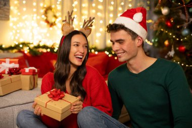 Noel için süslenmiş oturma odasında oturan romantik çift, hediye kutusunu tutan ve erkeğe gülümseyen mutlu kadın.