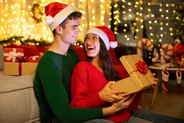 Şaşırmış Avrupalı çift sihirli Noel hediye kutusunu açıyor, Noel arifesinde ya da Noel arifesinde dekore edilmiş oturma odasında kış tatillerinin tadını çıkarıyorlar.