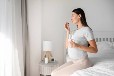 Hap alan ve elinde bir bardak su tutan genç bir kadın. Yatak odasında, boş bir yerde oturuyor. Hamilelik vitaminleri, takviyeler
