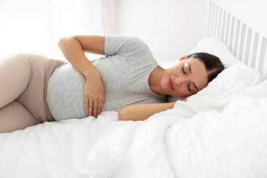 Sakin hamile kadın evde rahat bir yatakta uyuyor, karnına dokunuyor, tembelce boş vakitlerin tadını çıkarıyor, yumuşak yastıkta dinleniyor, temiz beyaz ketenler seriyor.