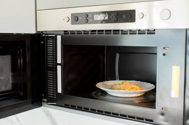 Beyaz kabında spagetti bulunan mikrodalga fırını açık, mutfakta beyaz rafta, yakın çekim.