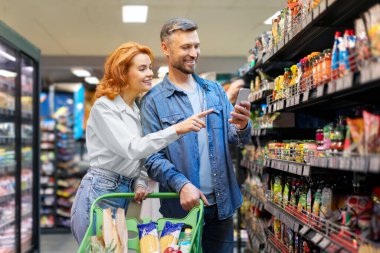 Avrupalı aile süpermarkette market alışverişi yapıyor, cep telefonu kullanıyor ve mağaza koridorunda duran ürünleri seçiyor.