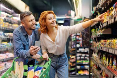 Çift birlikte market alışverişi yapıyor, erkek cep telefonu kullanıyor, kadın rafı işaret ediyor, süpermarketten yiyecek ürünleri seçiyor.