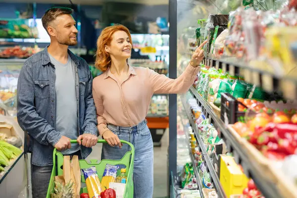 Süpermarkette yiyecek seçen aile çifti, market alışverişi, taze sebzeleri işaret eden kadın. Marketten ürün alan tüketiciler