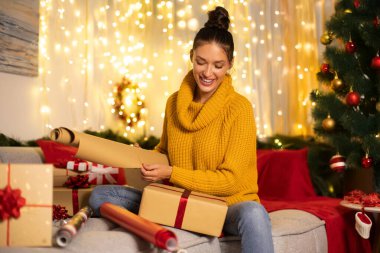 Mutlu Avrupalı kadın hediye paketlerini el sanatları kağıdıyla paketliyor, dekore edilmiş odada koltukta oturuyor ve yılbaşı ağacıyla süslüyor.