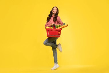 Sepet dolusu yiyecek ürünüyle poz veren uzun boylu bir kadın, sarı arka planda tek başına duruyor, stüdyo portresi.