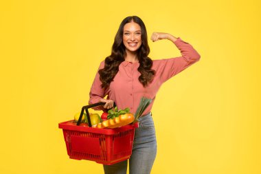 Kırmızı sepeti elinde yiyecek ürünleriyle tutan ve el pazısı kaslarını gösteren mutlu genç bayan sarı arka planda güç gösterisi yapıyor.