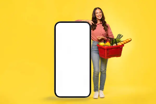 Süpermarket ya da gıda dağıtım uygulaması. Sepet dolusu ürünü olan mutlu kadın, sarı arka planda boş ekranı olan akıllı telefonun yanında duruyor.