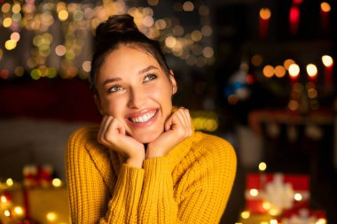 Noel ağacının önünde oturan hayalperest mutlu kadın ve akşamları Xmas ışıkları, yan bakıp gülümsüyor, Yeni Yıl Arifesi 'nin tadını çıkarıyor, bedava uzay.