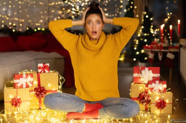 Kafasına dokunan ve kameraya bakan, kötü haberler duyan, hediye kutuları ve çelenk ışıklarıyla süslenmiş bir odada oturan genç bayan.