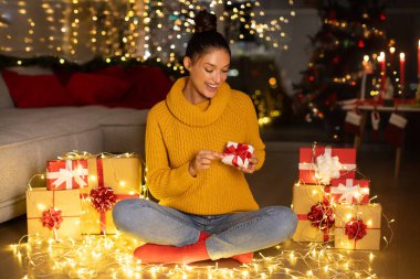 Mutlu kadın kucağında ve küçük bir Noel hediyesi kutusu açıyor, hediye kutularının ve evdeki çelenklerin arasında yerde oturuyor.