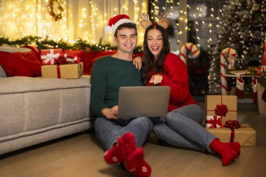 Mutlu eşler dizüstü bilgisayar kullanır, Noel hediyelerini seçer, ekranı işaret eder, dekore edilmiş oturma odasında Noel ağacı ve çelenklerle oturur.