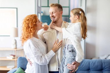 Üç kişilik Avrupalı aile, anne, baba ve kız kucaklaşırken oturma odasında iç içe geçiyor, kaynaşıyor ve gülümsüyor.