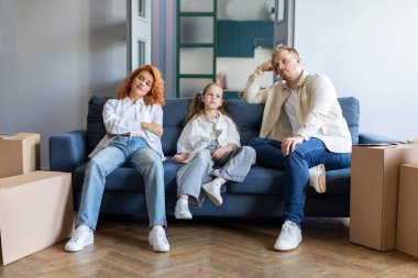 Üç kişilik üzgün bir aile. Oturma odasındaki kanepede oturuyor. Yanında da eşyalarıyla birlikte karton kutular var.