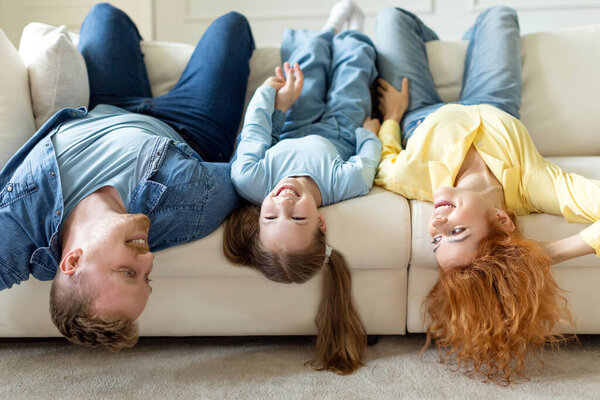 Family of three fooling around lying on sofa upside down and laughing, parents with daughter having fun on cozy couch