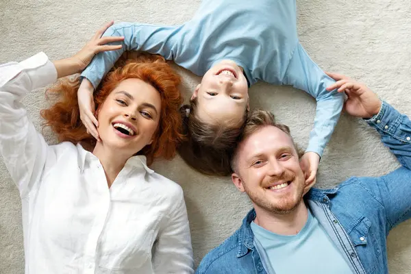 Üç kişilik mutlu bir aile halı üzerinde uzanıyor ve birbirlerine bağlanıyorlar, kameraya neşeyle gülümsüyorlar, baba, anne ve kız sarılıyorlar, üst manzara
