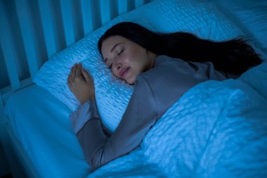 Güzel genç bir kadın geceleri yatağında uyuyor, battaniyenin altında yatıyor ve gülümsüyor, rahatlığın tadını çıkarıyor ve yatak odasında dinleniyor.