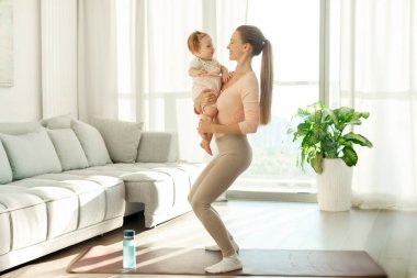 Bebekle spor aktiviteleri. Anne, kızıyla evde egzersiz yapıyor, kızını kollarına alıyor ve çömeliyor, spor paspası üzerinde çalışıyor.