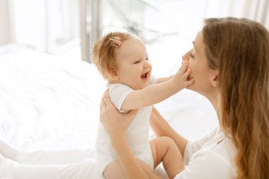 Genç anne evde kızıyla oynuyor, yatak odasında oturuyor, çocuk annesinin yüzüne dokunuyor ve gülümsüyor, yan manzara, boş alan.