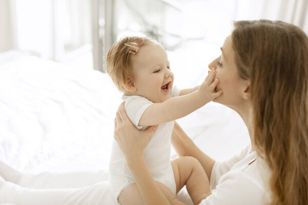 Young mother playing with her baby girl at home, sitting in the bedroom, kid touching moms face and smiling, side view, free space